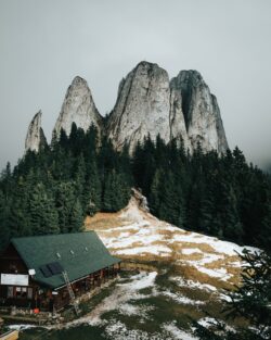 mountain cabin towering rocks pine forest rocky peaks alpine house hiking shelter autumn snow wilderness landscape dramatic mountains remote dwelling evergreen trees misty weather rocky spire natio
