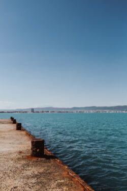 rusty pier concrete dock turquoise sea blue sky coastal town distant buildings industrial structure calm water sunny day worn texture waterfront ocean horizon maritime Mediterranean coast seascape